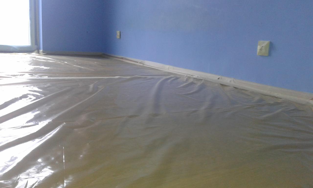 Pokryta folią ochronną podłoga w pokoju z pomalowanymi na niebiesko ścianami, zabezpieczone listwy przypodłogowe i gniazdko elektryczne.