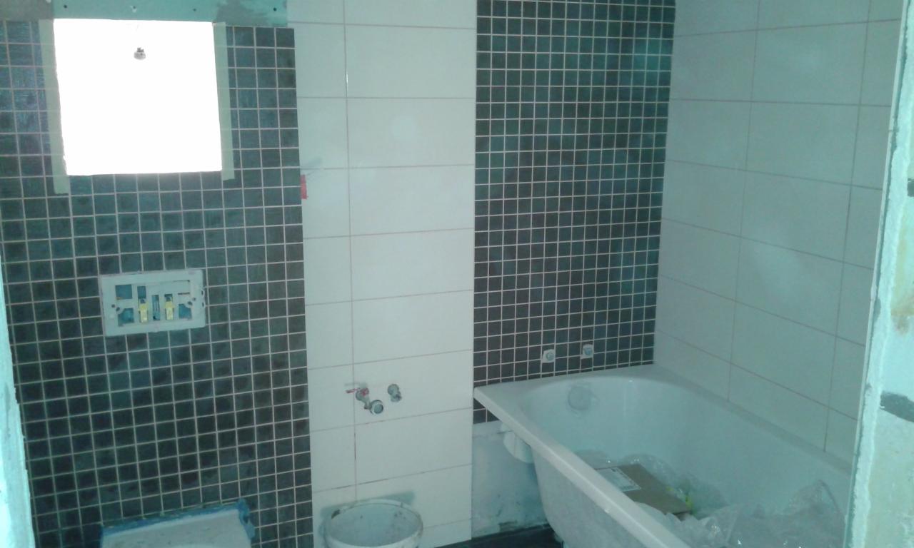 Łazienka w trakcie wykańczania: częściowo ułożone płytki w kolorze grafitowym i białym, widoczna wanna, puszka elektryczna i wiadro z zaprawą.