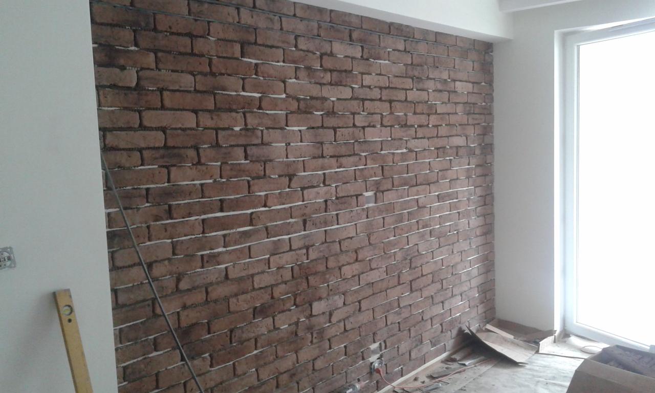 Ściana z cegły dekoracyjnej w trakcie montażu, widoczne narzędzia i materiały budowlane, poziomica przy ścianie, okno w tle.