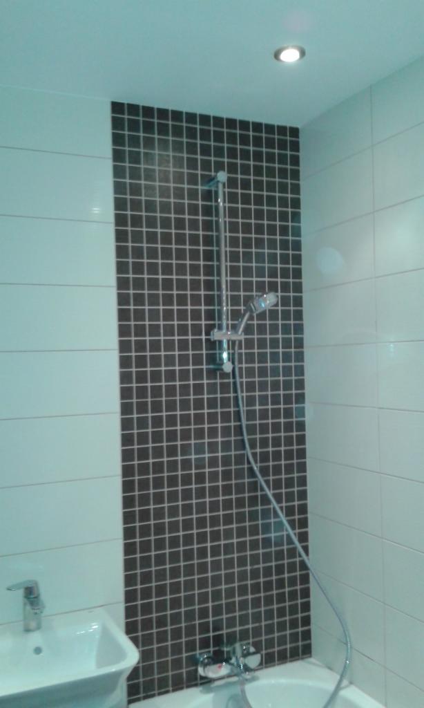 Wykończona łazienka z białymi płytkami i pionowym pasem ciemnej mozaiki za prysznicem, widoczna bateria umywalkowa i wannowa.