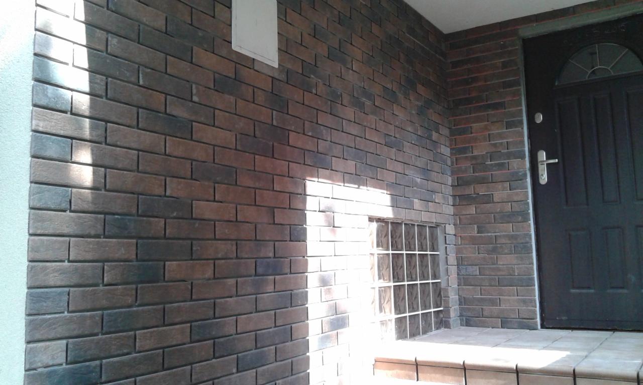 Balkon wykończony klinkierem w odcieniach brązu i grafitu, z małym okienkiem z luksferów i czarnymi drzwiami wejściowymi. Na ścianie widoczny prostokątny element wentylacyjny.