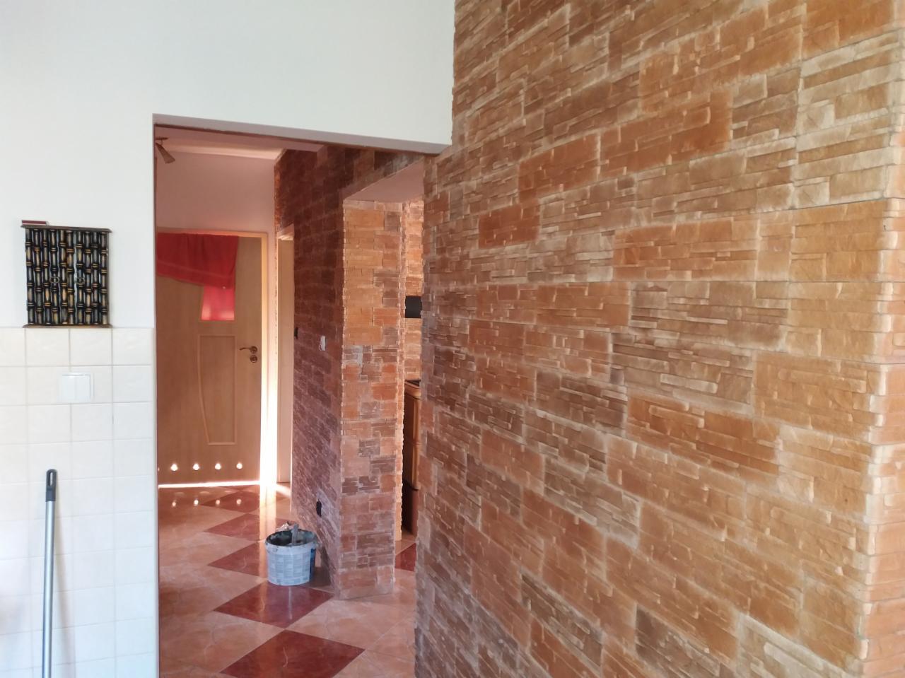 Ściana wykończona dekoracyjną cegłą w odcieniach brązu, widok z korytarza na pokój z czerwoną zasłoną i wzorzystą podłogą, wiadro z farbą przy ścianie.