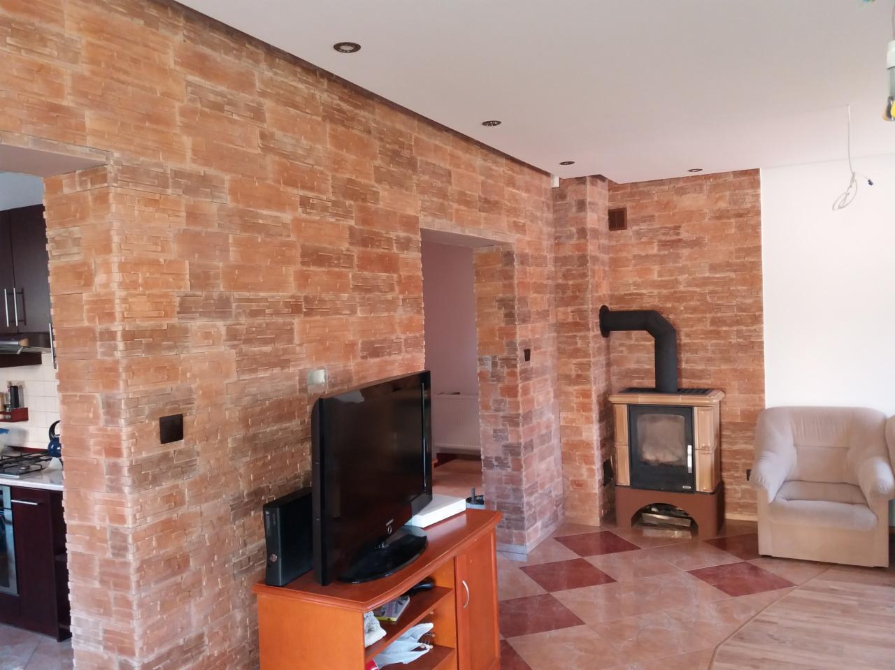 Salon z ceglaną ścianą, kominkiem kaflowym z czarną rurą i telewizorem na drewnianej komodzie, widoczny fragment kuchni w tle.