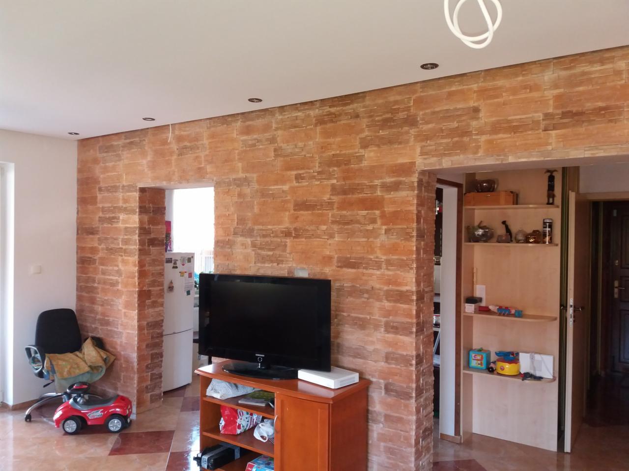 Salon z dekoracyjną ścianą z cegły, telewizorem na komodzie i widokiem na otwarte drzwi do kuchni i pokoju z regałem.