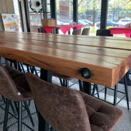 Długi, drewniany blat barowy z ciemnymi, skórzanymi krzesłami barowymi na metalowych nogach, widok z perspektywy siedzącego klienta, jasne wnętrze lokalu z widokiem na ulicę.