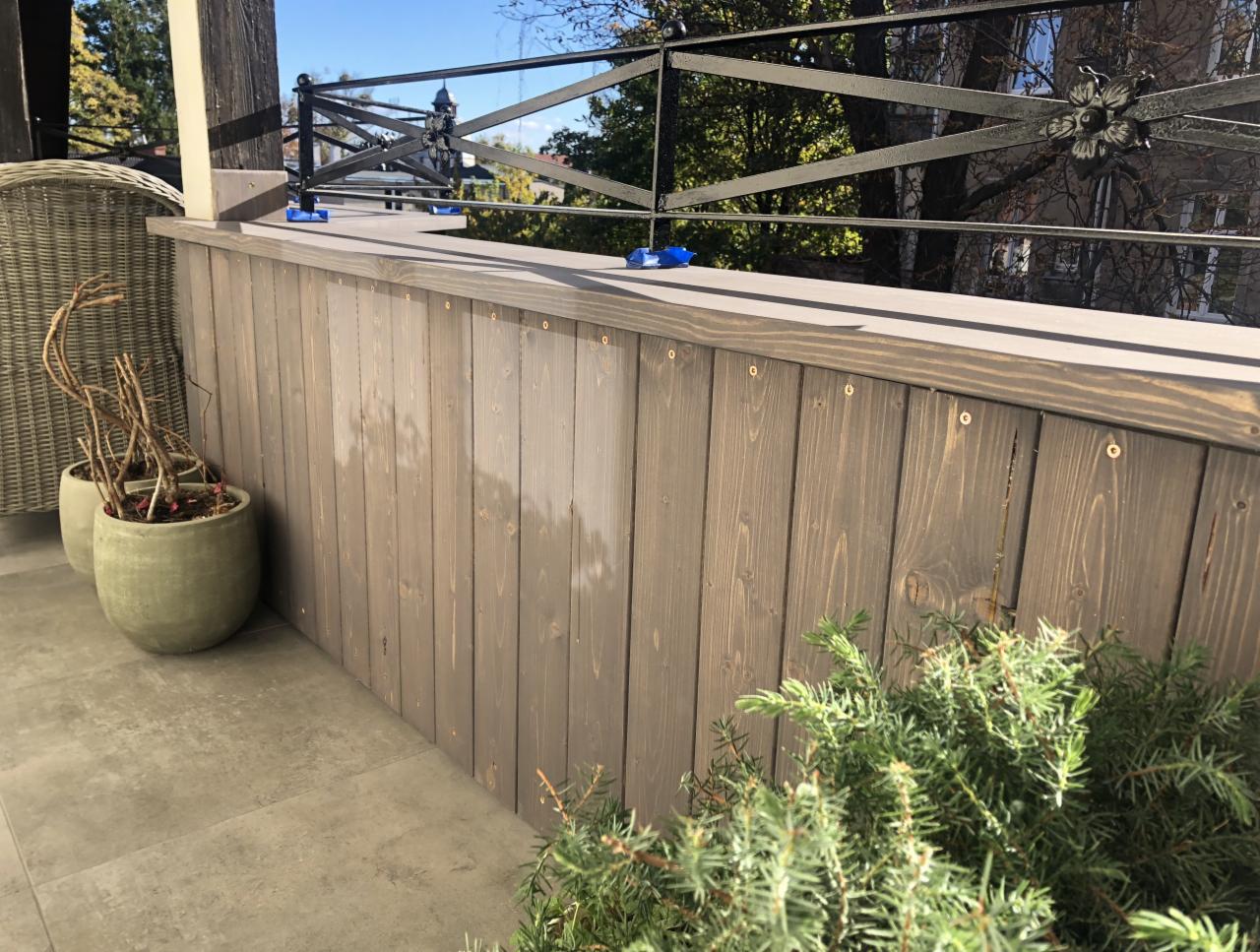 Drewniana balustrada balkonowa w szarym odcieniu, z kutą, czarną poręczą i roślinami w donicach na szarym balkonie.