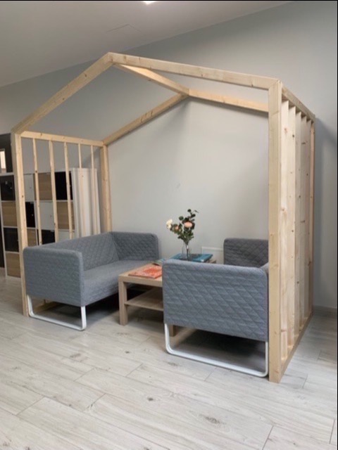 Szara sofa i fotel z pikowanym obiciem, umieszczone w drewnianej konstrukcji przypominającej domek, na podłodze imitującej drewno.