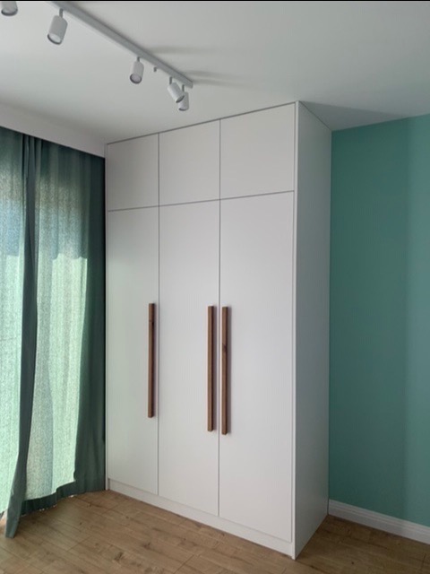 Biała, wysoka szafa z drewnianymi uchwytami, ustawiona w rogu pokoju z turkusową ścianą i parkietem, oświetlona szynoprzewodem.