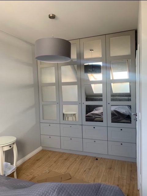 Szara zabudowa szaf z lustrzanymi frontami i szufladami w sypialni na poddaszu z widocznym skosem dachu, oświetlona wiszącą lampą z abażurem w tym samym odcieniu szarości.
