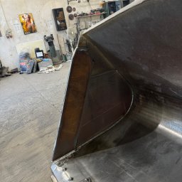 Serwis spawalniczy Piotr Kaźmierczak - Metalowa łyżka koparki po naprawie, widoczne spawy i mocowania, w tle warsztat z narzędziami i plakatem na ścianie.