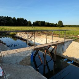 Serwis spawalniczy Piotr Kaźmierczak - Metalowa kładka nad betonowym kanałem wodnym na tle zielonego pola i drzew. Widoczne ślady prac budowlanych, worki i taśmy ostrzegawcze.