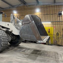 Serwis spawalniczy Piotr Kaźmierczak - Szara ładowarka Volvo z metalową łyżką w warsztacie. Widoczne ślady spawania na łyżce i masywne opony ładowarki. W tle ściana z drewnianą boazerią i żółtym pasem.