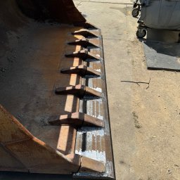 Serwis spawalniczy Piotr Kaźmierczak - Wytrzymała łyżka koparki z zębami po spawaniu, widoczne ślady obróbki. Na betonie leży drut. W tle metalowe kontenery i ogrodzenie z siatki.