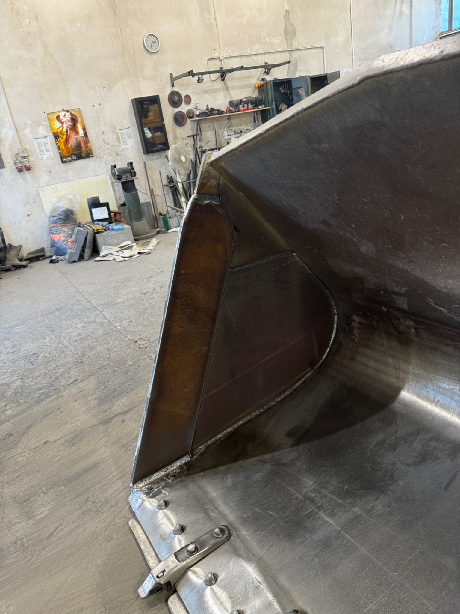 Metalowa łyżka koparki po naprawie, widoczne spawy i mocowania, w tle warsztat z narzędziami i plakatem na ścianie.