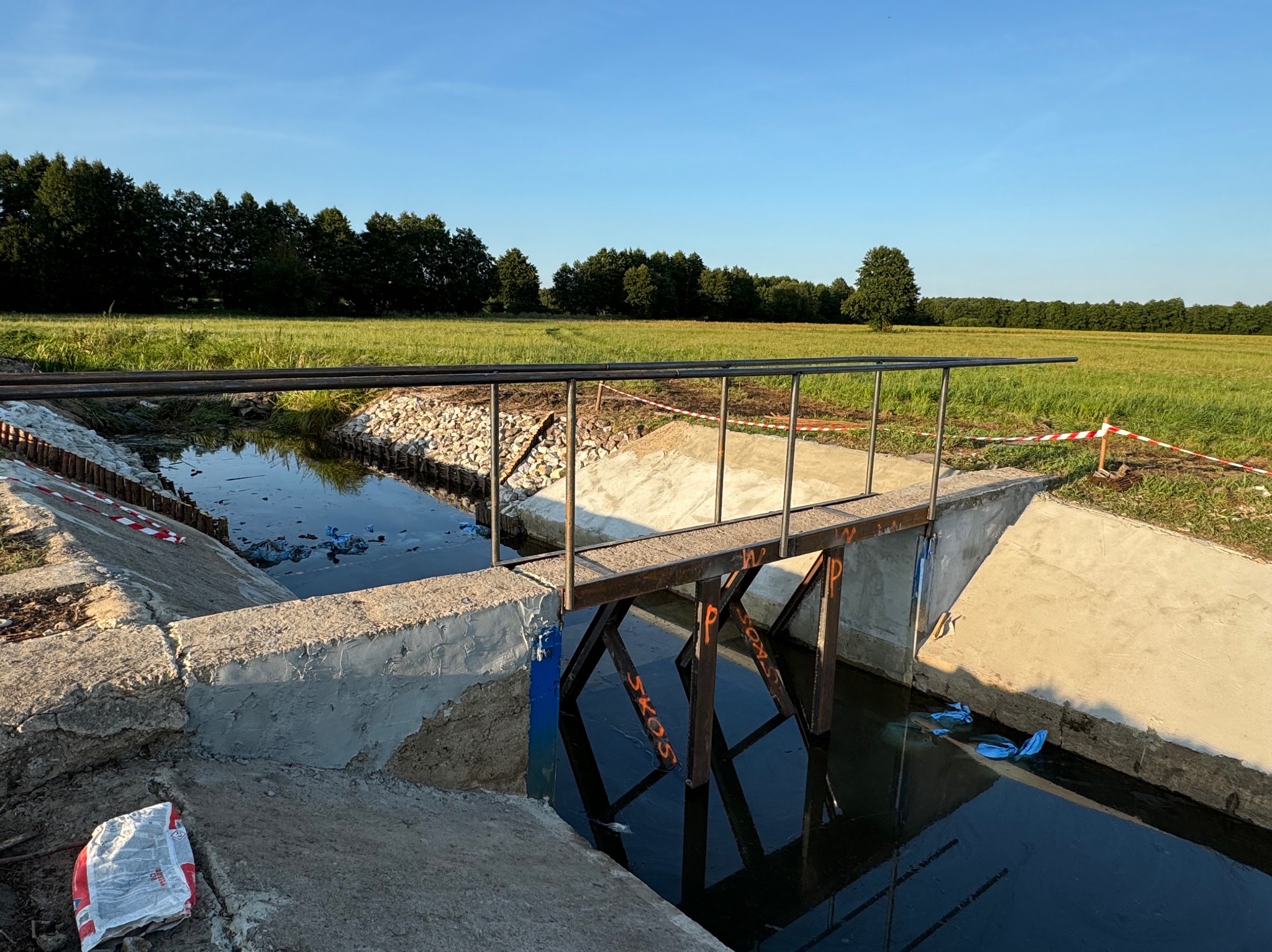 Metalowa kładka nad betonowym kanałem wodnym na tle zielonego pola i drzew. Widoczne ślady prac budowlanych, worki i taśmy ostrzegawcze.