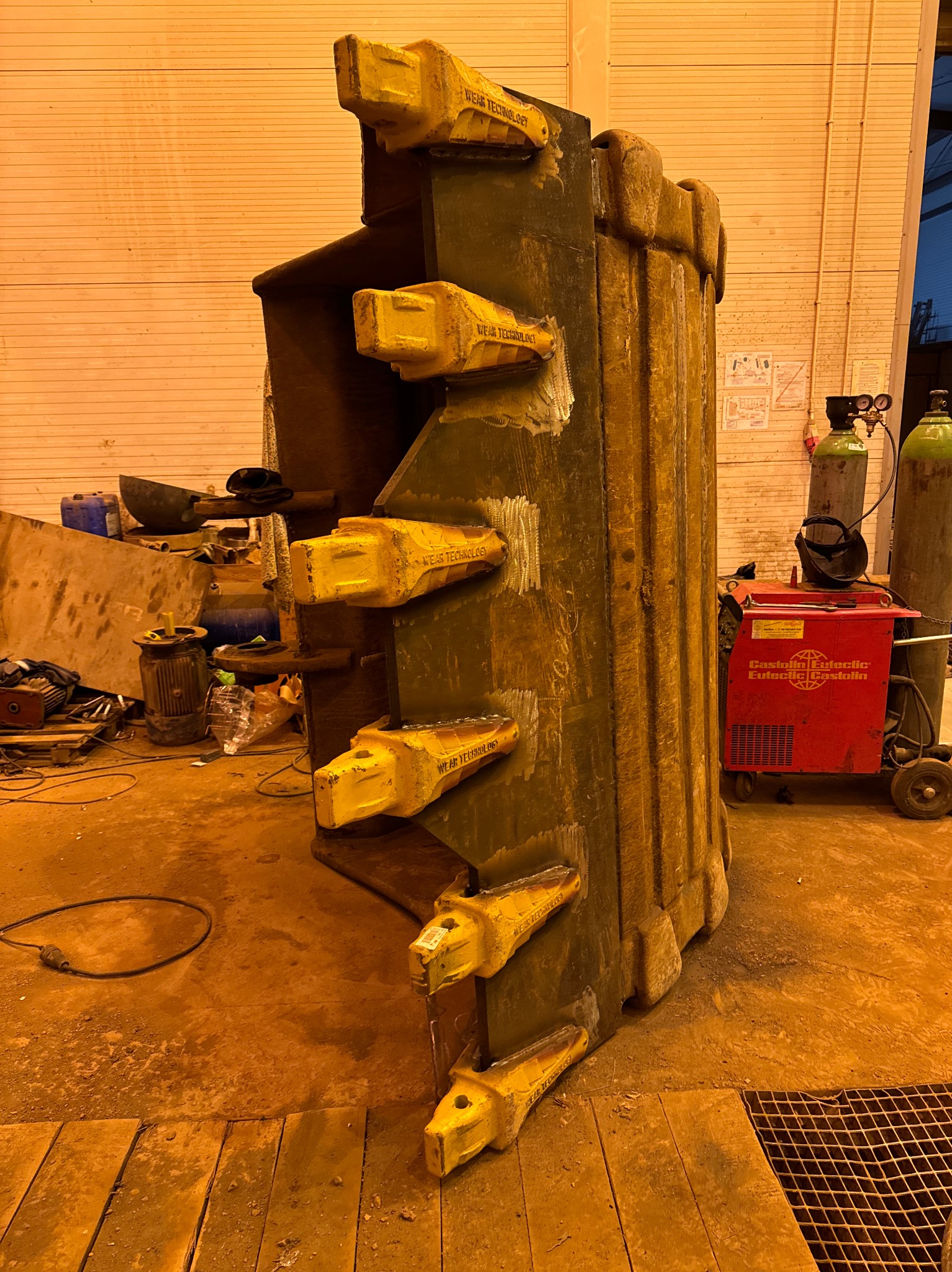 Ciężka, spawana konstrukcja stalowa z żółtymi elementami wymiennymi, prawdopodobnie z maszyny budowlanej, widoczne ślady spawania, w tle butle z gazem i czerwony agregat spawalniczy Castolin Eutectic.