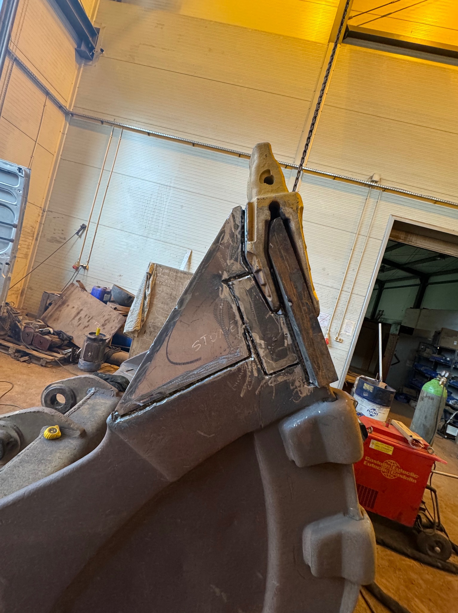 Ciężki element metalowy, prawdopodobnie łyżka koparki, po spawaniu w warsztacie, widoczne nowe spawy i napis 'STUC' na boku, w tle spawarka i butla z gazem.