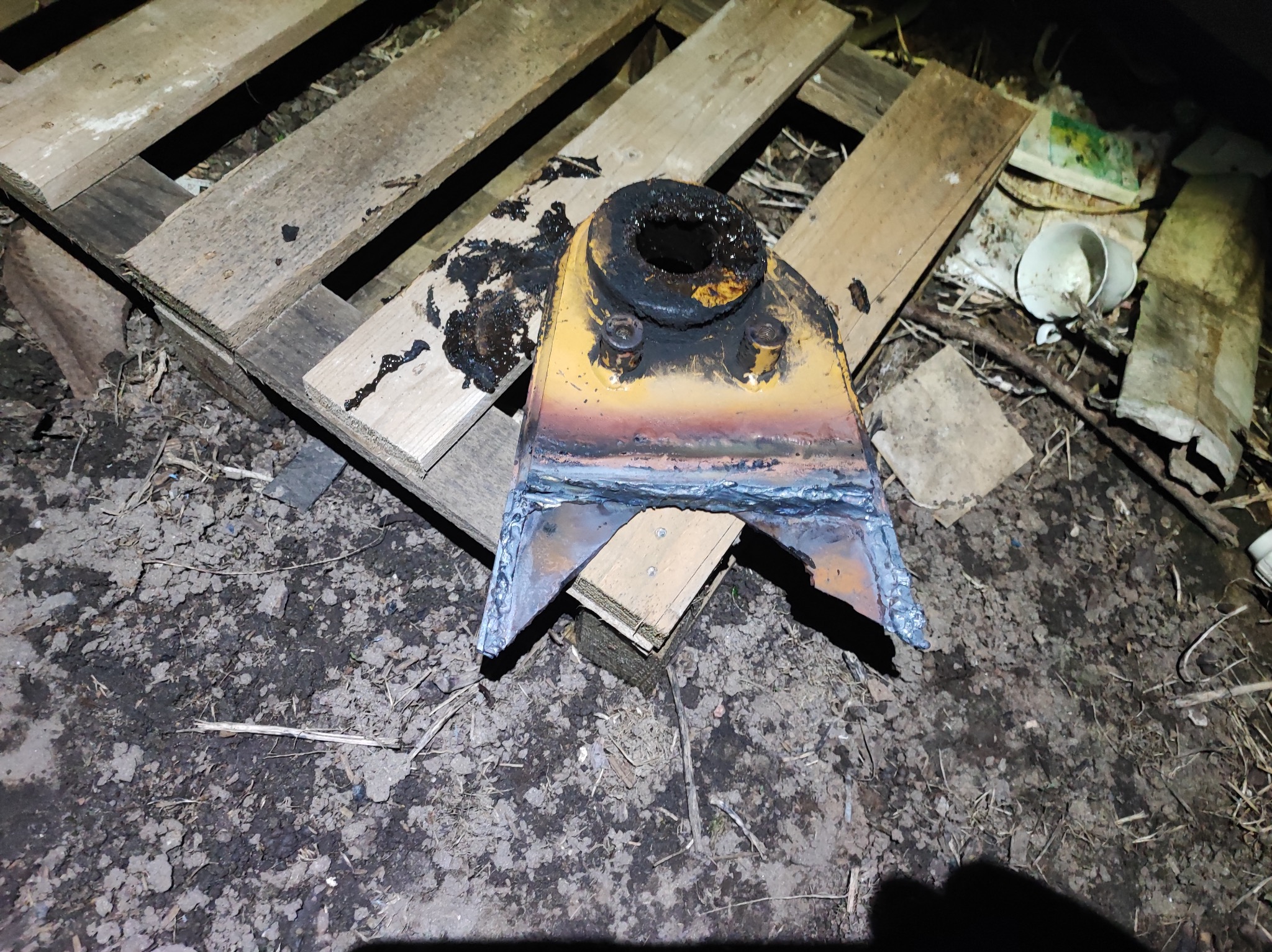 Żółta, metalowa konstrukcja z widocznymi śladami spawania, leżąca na drewnianej palecie na tle ziemi i śmieci.