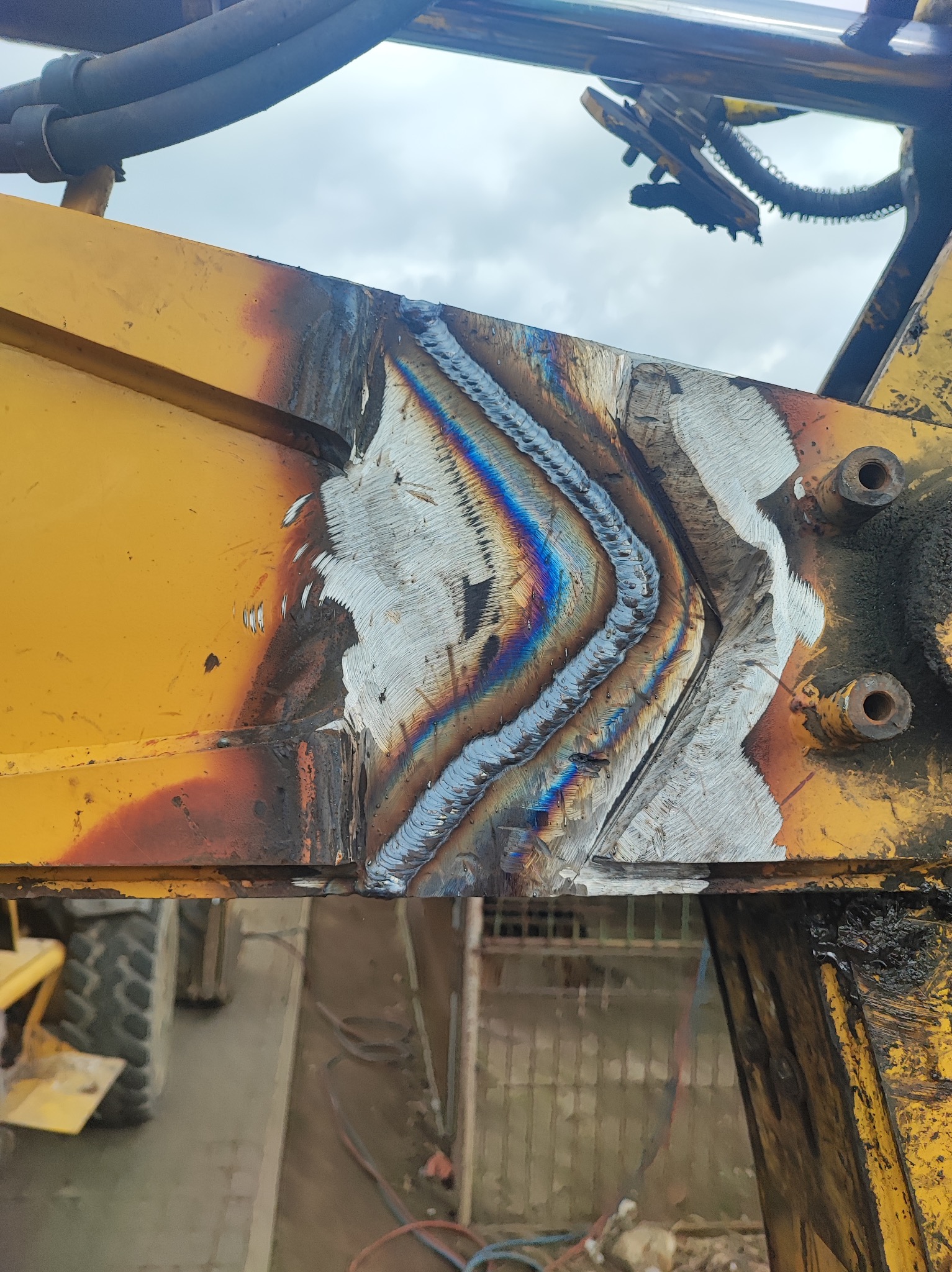 Żółty element konstrukcyjny maszyny budowlanej po spawaniu, widoczne tęczowe przebarwienia metalu wzdłuż spoiny.