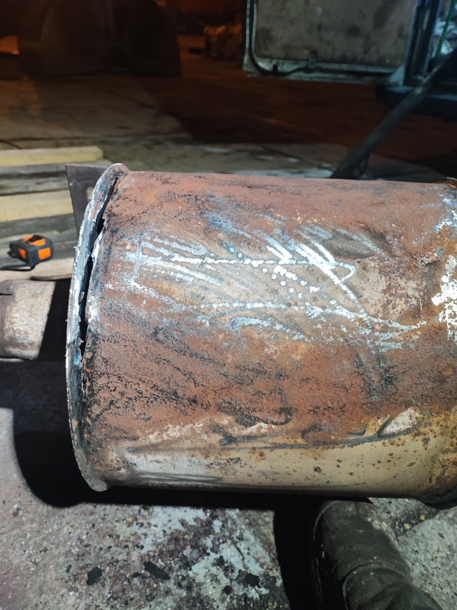 Zardzewiały, cylindryczny element metalowy z widocznymi śladami spawania i cięcia, leżący na ziemi, obok miarki zwijanej.