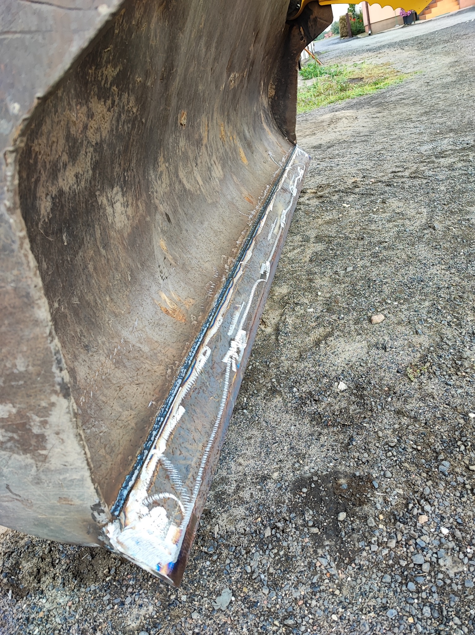 Metalowa łyżka koparki z widocznymi śladami spawania na krawędzi, leżąca na żwirowym podłożu.