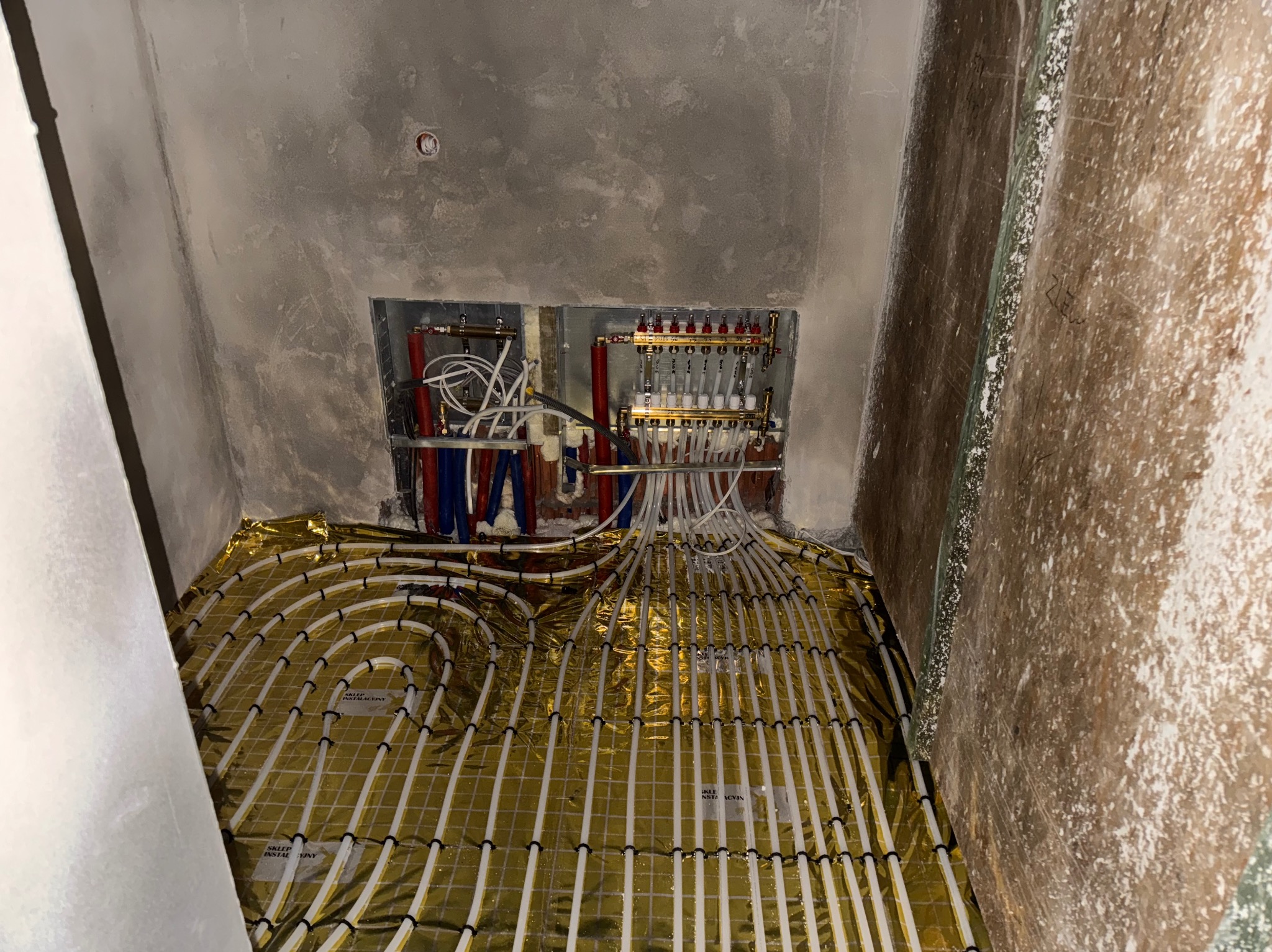 Instalacja ogrzewania podłogowego w trakcie montażu: białe rurki ułożone na złotej folii izolacyjnej, widoczne rozdzielacze na ścianie w surowym stanie.