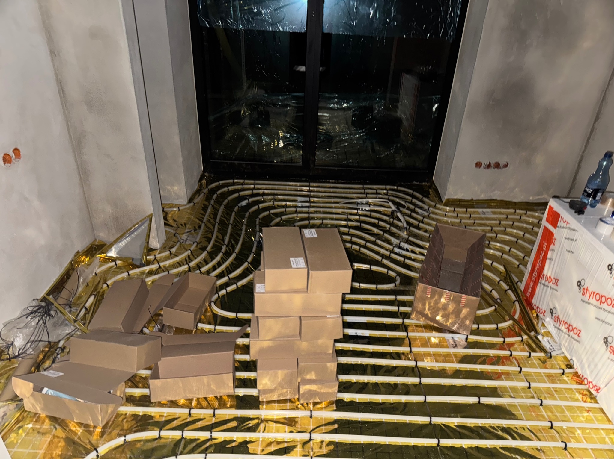 Ułożone rury ogrzewania podłogowego na folii izolacyjnej w pomieszczeniu z dużym oknem, przygotowane do wylania posadzki, z widocznymi kartonami i materiałami budowlanymi.