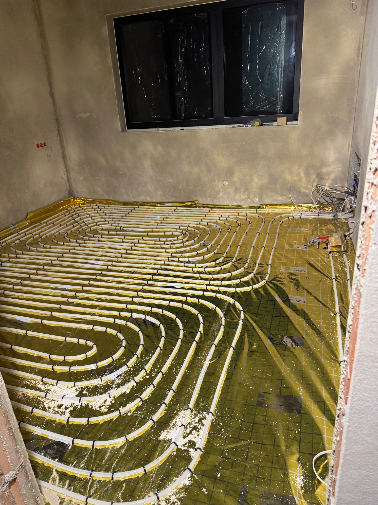 Białe rury ogrzewania podłogowego ułożone spiralnie na złotej folii izolacyjnej w surowym wnętrzu, widok z góry pod kątem. Widoczne narzędzia i okno.