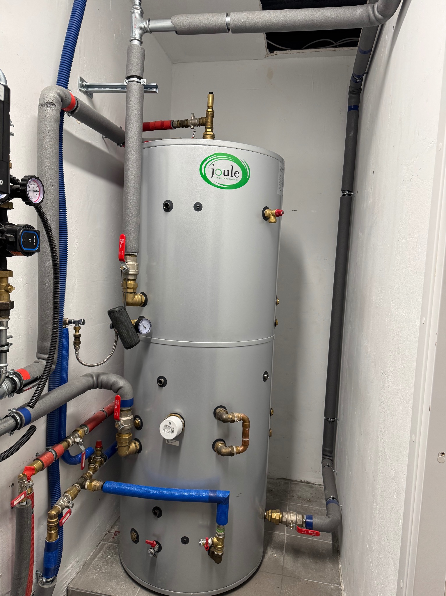 Szary, pionowy zbiornik Joule w kotłowni, z licznymi rurami i zaworami. Instalacja wodna w piwnicy, z izolowanymi rurami i manometrami.