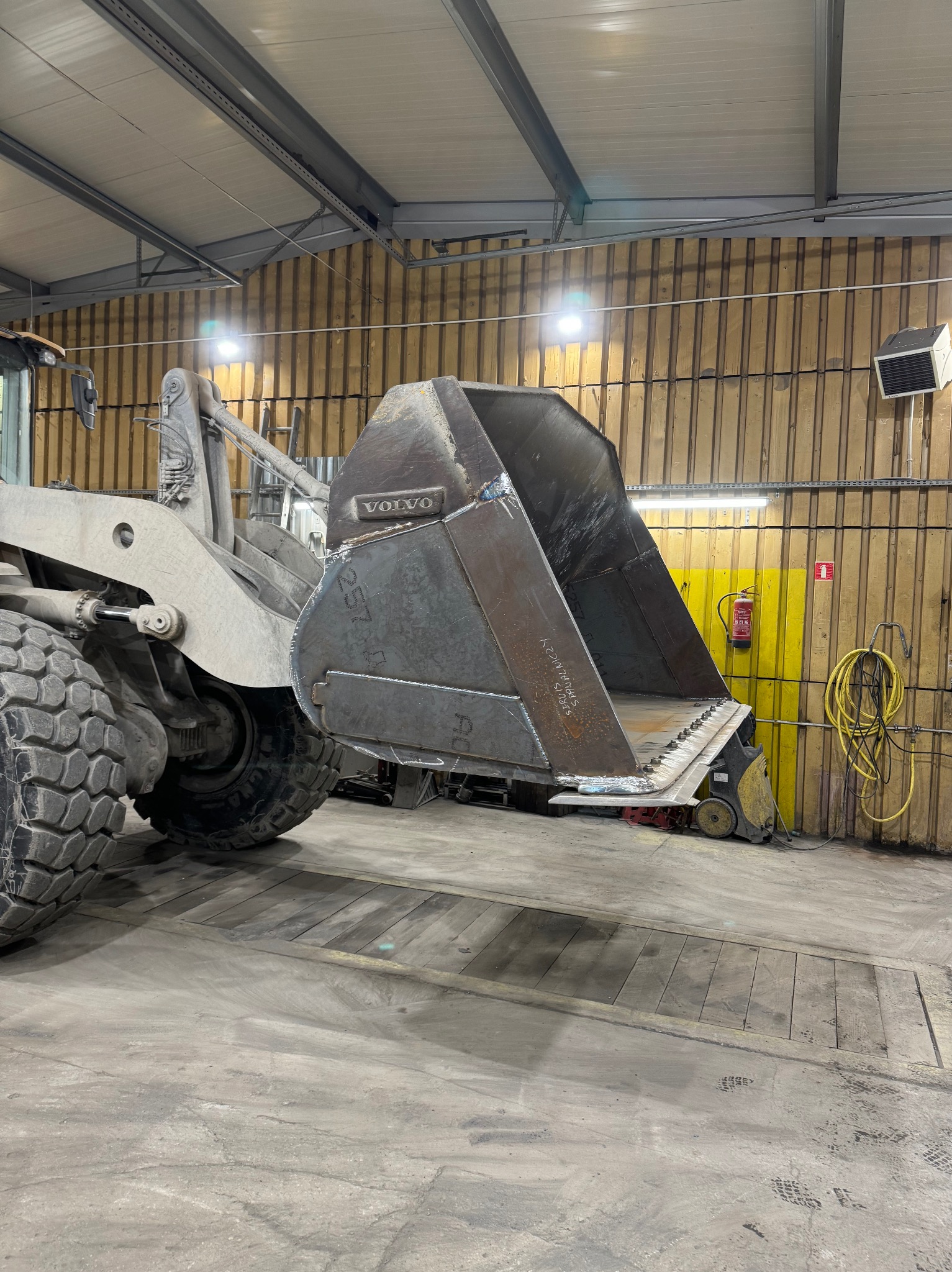 Szara ładowarka Volvo z metalową łyżką w warsztacie. Widoczne ślady spawania na łyżce i masywne opony ładowarki. W tle ściana z drewnianą boazerią i żółtym pasem.