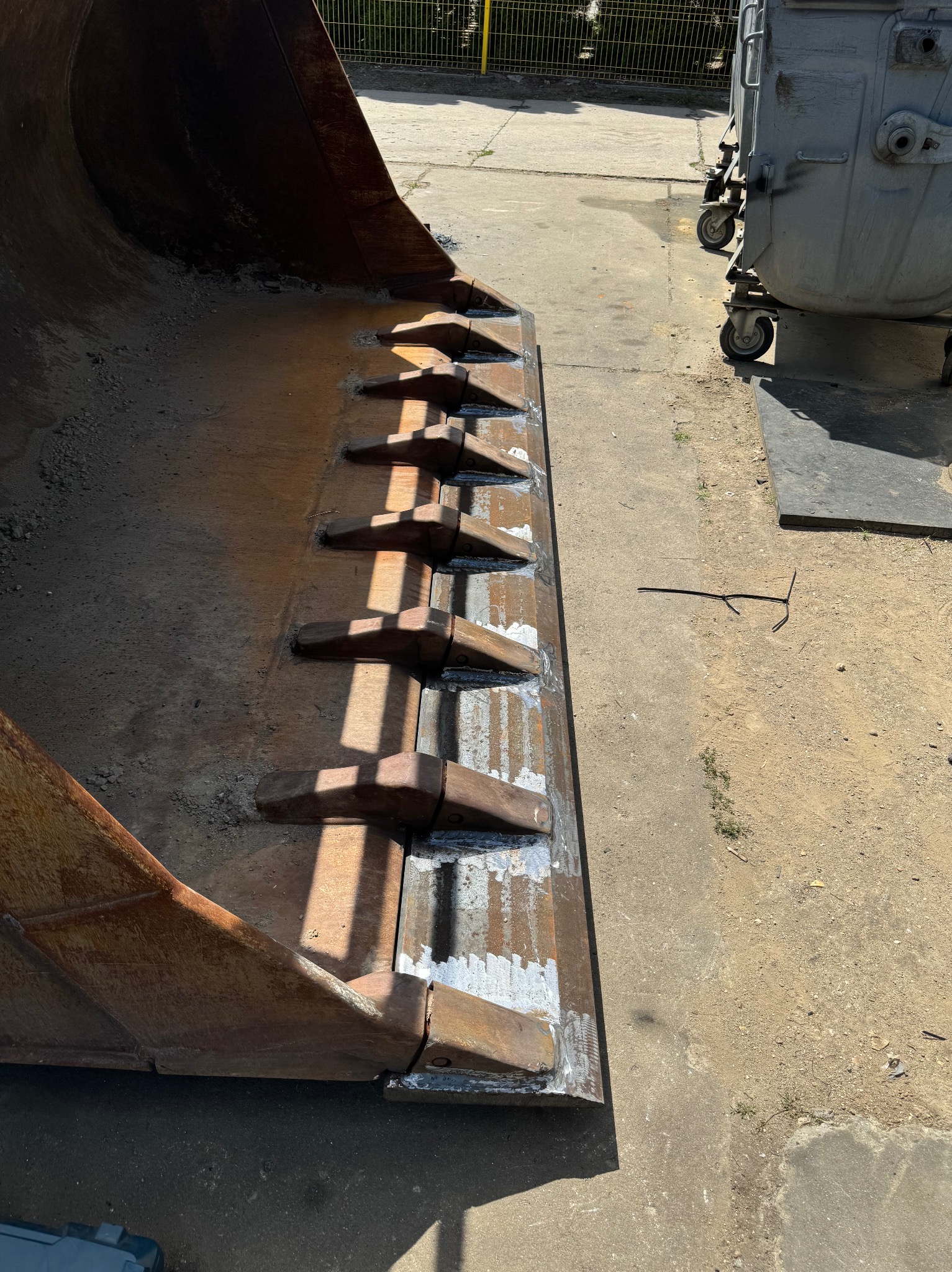 Wytrzymała łyżka koparki z zębami po spawaniu, widoczne ślady obróbki. Na betonie leży drut. W tle metalowe kontenery i ogrodzenie z siatki.