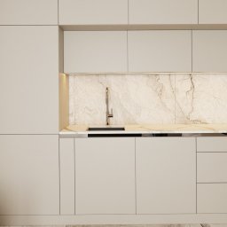de AR - Minimalistyczna kuchnia z beżowymi, gładkimi frontami szafek, marmurowym blatem i backsplashiem, oświetlona subtelnym światłem LED pod szafkami wiszącymi, z widocznym fragmentem marmurowej podłogi.