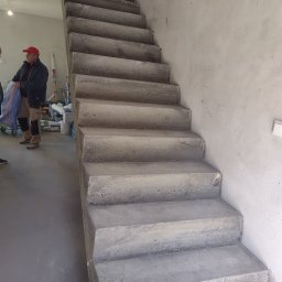 SPRINT-BUD Zbigniew Koloch - Surowe, betonowe schody wewnętrzne w trakcie budowy, widoczne ślady po szalunku, robotnik w czerwonej czapce w tle.