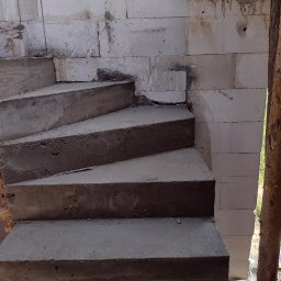 SPRINT-BUD Zbigniew Koloch - Surowe, betonowe schody spiralne w trakcie budowy, przylegające do ściany z bloczków betonowych, z widocznym drewnianym szalunkiem i fragmentem krajobrazu w tle.