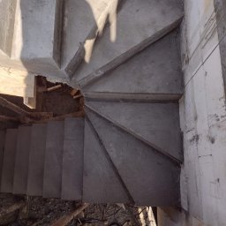 SPRINT-BUD Zbigniew Koloch - Surowe, betonowe schody spiralne w trakcie budowy, widok z góry z widocznym cieniem osoby, szalunki i gruz pod schodami.