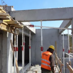 Budowa domu murowanego: betonowe ściany i strop w trakcie wznoszenia, widoczne podpory i szalunki, pracownik w odblaskowej kamizelce i kasku na tle zieleni.