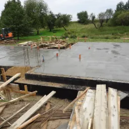 Wylany fundament pod budowę z widoczną izolacją, wystającymi prętami zbrojeniowymi, pomarańczowymi rurami instalacji i koparką w tle.