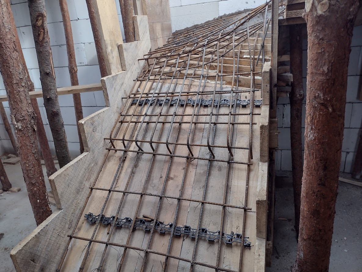Szkielet zbrojenia schodów z drewnianym szalunkiem i podporami z surowych pni drzew, widok z góry na konstrukcję w trakcie budowy.