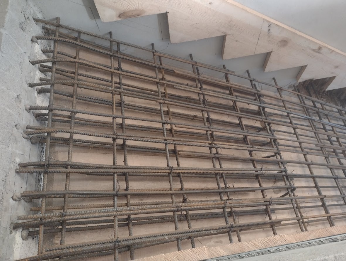 Siatka zbrojeniowa do betonowych schodów, widoczne drewniane szalunki i fragment surowego muru.