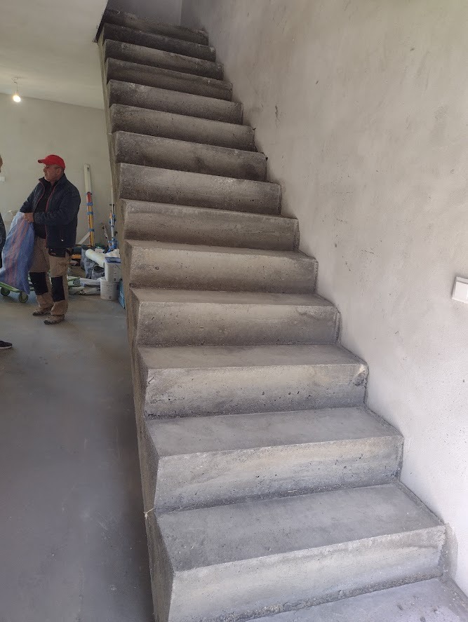 Surowe, betonowe schody wewnętrzne w trakcie budowy, widoczne ślady po szalunku, robotnik w czerwonej czapce w tle.