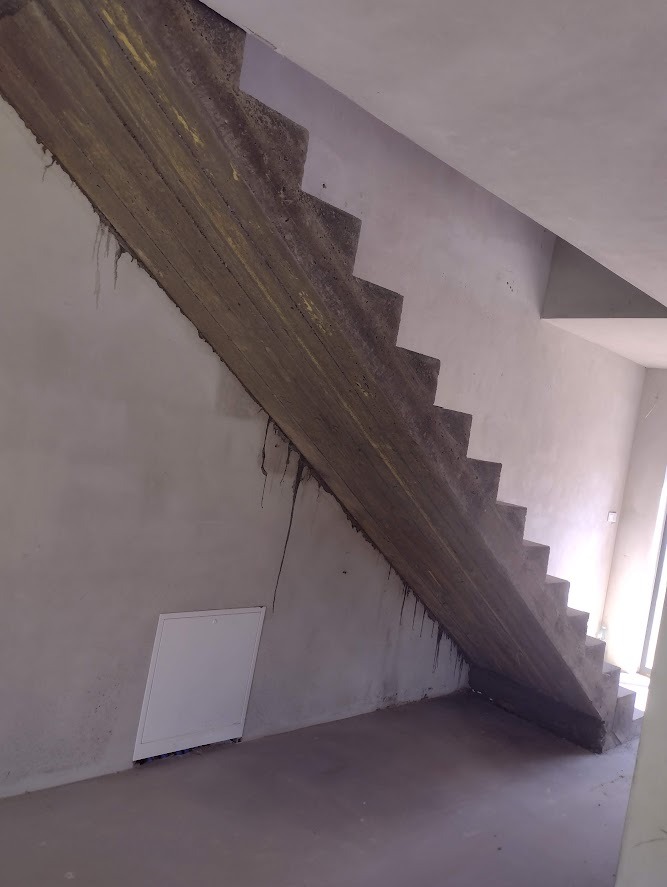 Surowe, betonowe schody wewnętrzne w trakcie budowy, widoczne ślady po zalewaniu betonu, biała klapa rewizyjna w ścianie.