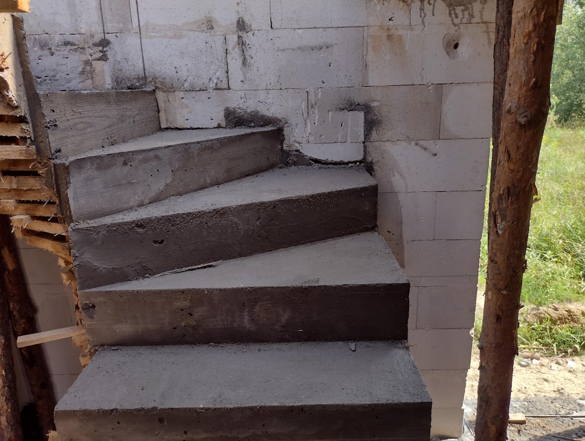 Surowe, betonowe schody spiralne w trakcie budowy, przylegające do ściany z bloczków betonowych, z widocznym drewnianym szalunkiem i fragmentem krajobrazu w tle.