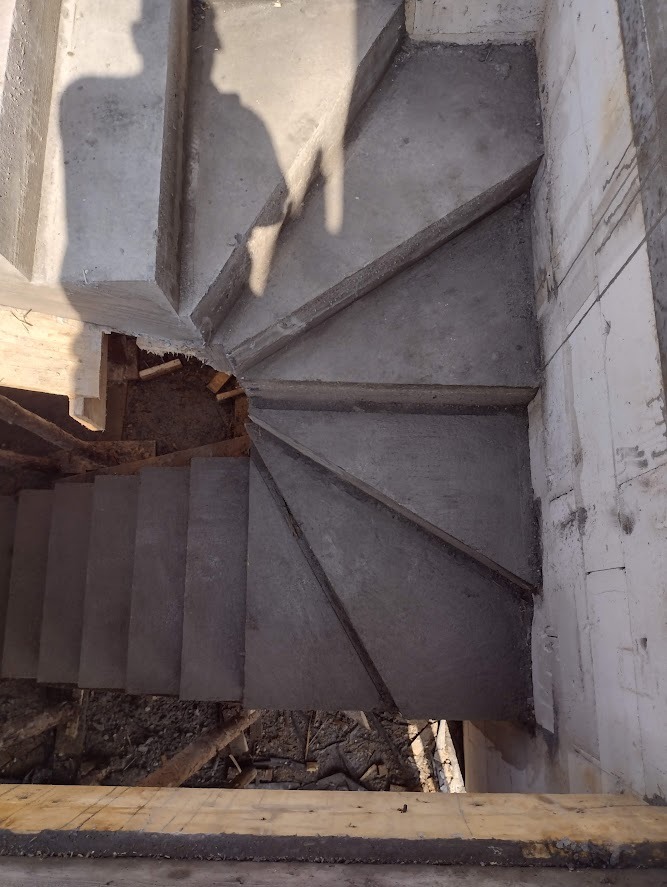 Surowe, betonowe schody spiralne w trakcie budowy, widok z góry z widocznym cieniem osoby, szalunki i gruz pod schodami.