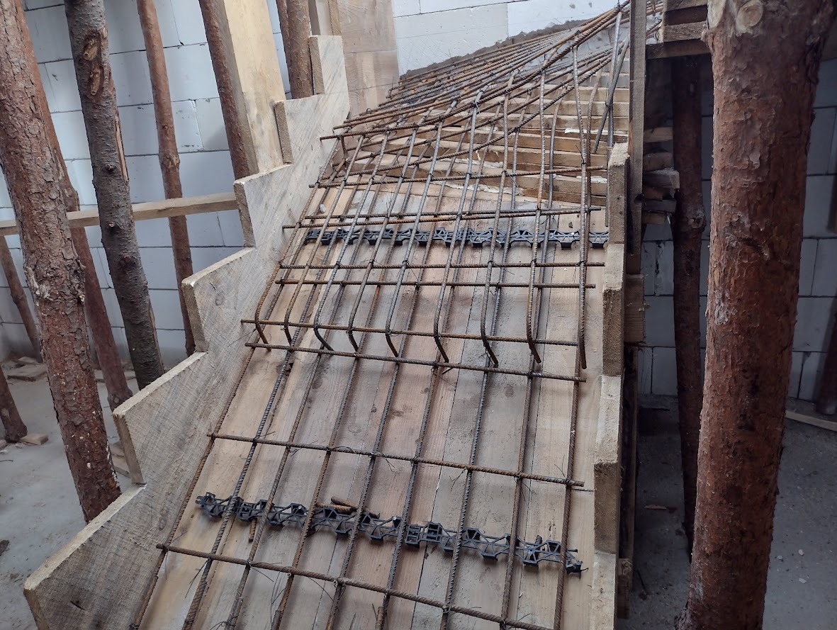 Szczegółowy widok szalunku drewnianego i zbrojenia stalowego przygotowanego pod wylewkę stopni schodów betonowych, wspartego na drewnianych stemplach.