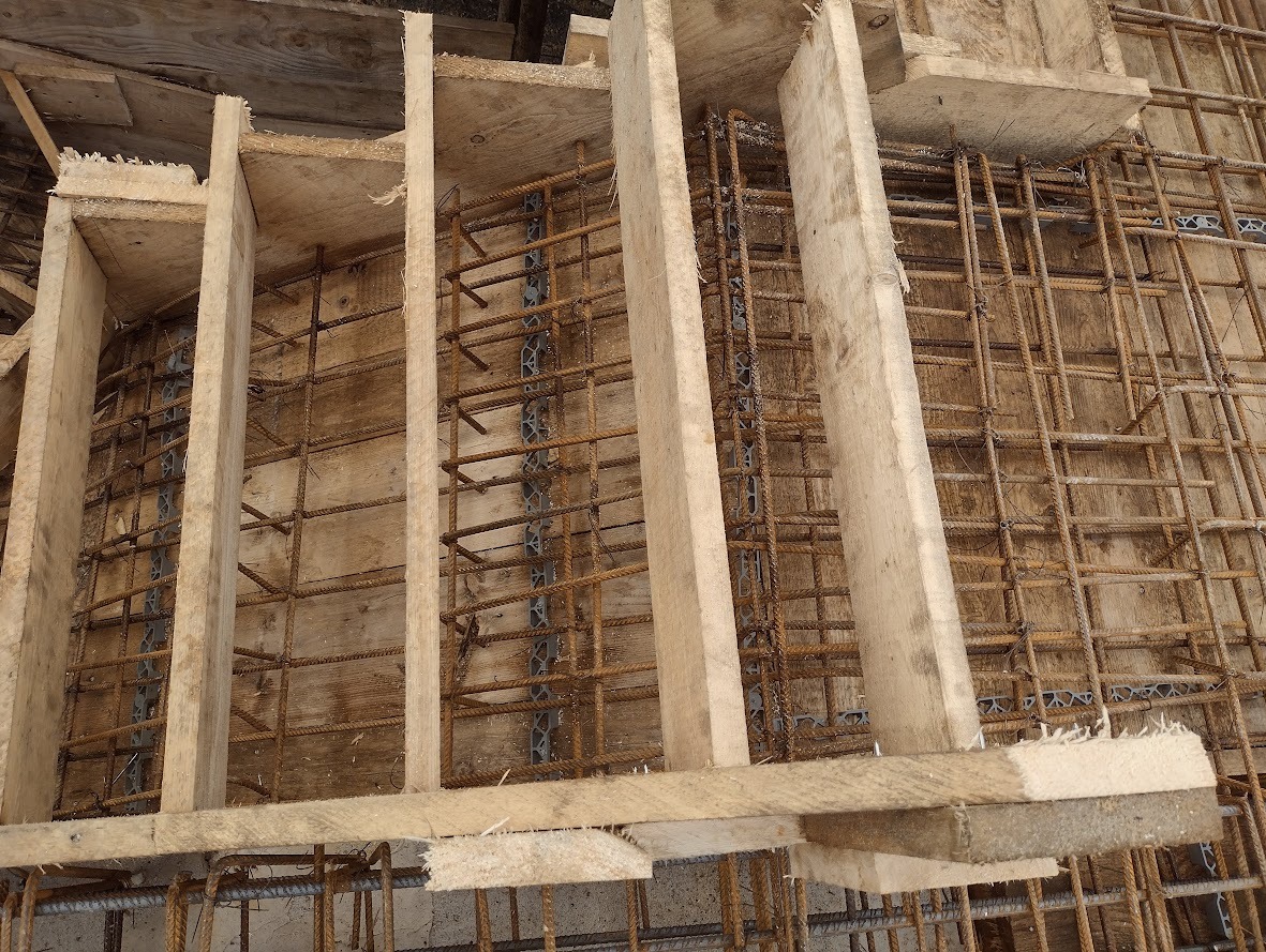 Szczegółowe ujęcie szalunku z desek i zbrojenia z prętów stalowych, tworzących siatkę zbrojeniową, połączonych plastikowymi dystansami, przygotowanych do zalania betonem.