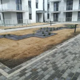Nowo założony ogród przed nowoczesnym budynkiem mieszkalnym z balkonami, widoczne rozłożone maty szkółkarskie i posadzone młode rośliny, obok ułożona szara kostka brukowa.