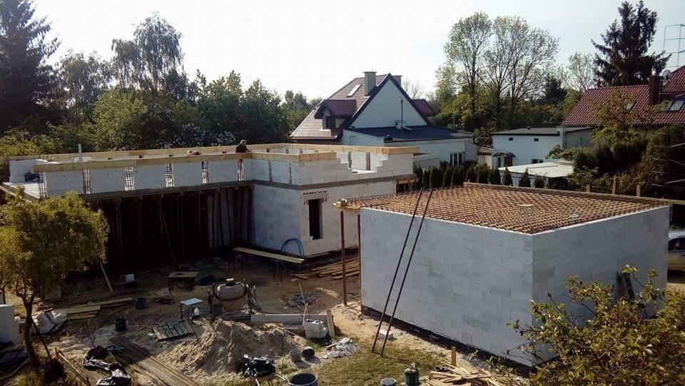 Budowa domu jednorodzinnego w trakcie realizacji, widoczne ściany z betonu komórkowego, szalunki, zbrojenie stropu, w tle dom mieszkalny i drzewa.