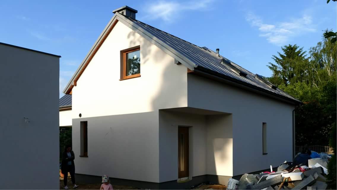 Nowoczesny dom jednorodzinny z ciemnoszarą blachodachówką, białymi ścianami i drewnianą stolarką okienną, widok z zewnątrz, z fragmentem drugiego budynku i elementami porządkowymi na pierwszym planie.