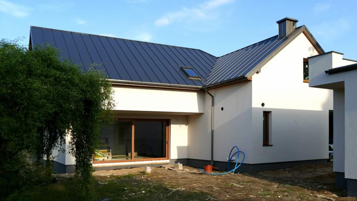 Nowoczesny dom jednorodzinny z antracytowym dachem, widok z zewnątrz na elewację i okno przesuwne, z niezagospodarowanym terenem wokół i niebieskim wężem ogrodowym.