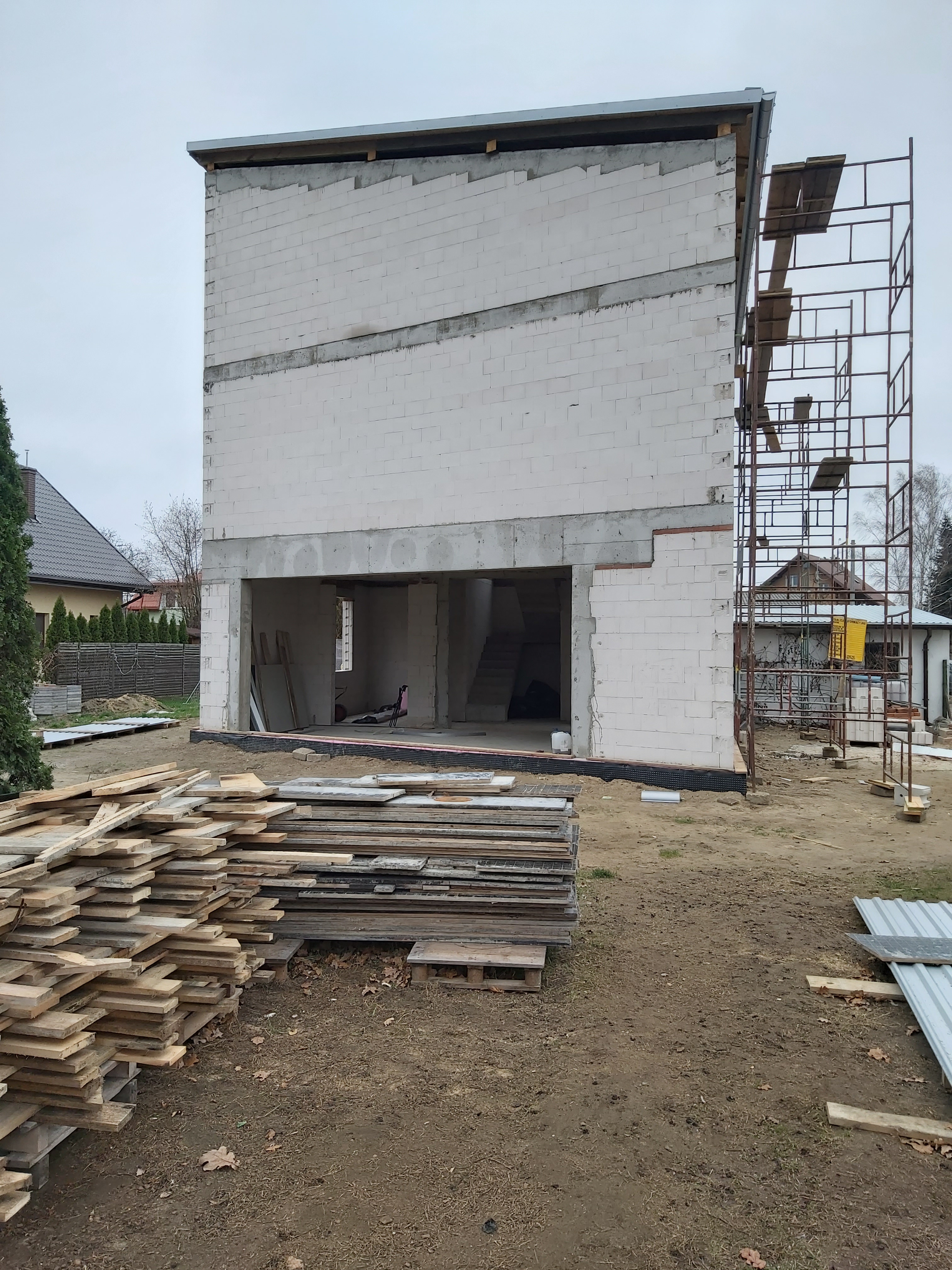 Surowa bryła budynku usługowego w trakcie budowy: ściany z bloczków betonowych, odsłonięte otwory okienne i drzwiowe, stos desek na pierwszym planie, rusztowanie z boku.